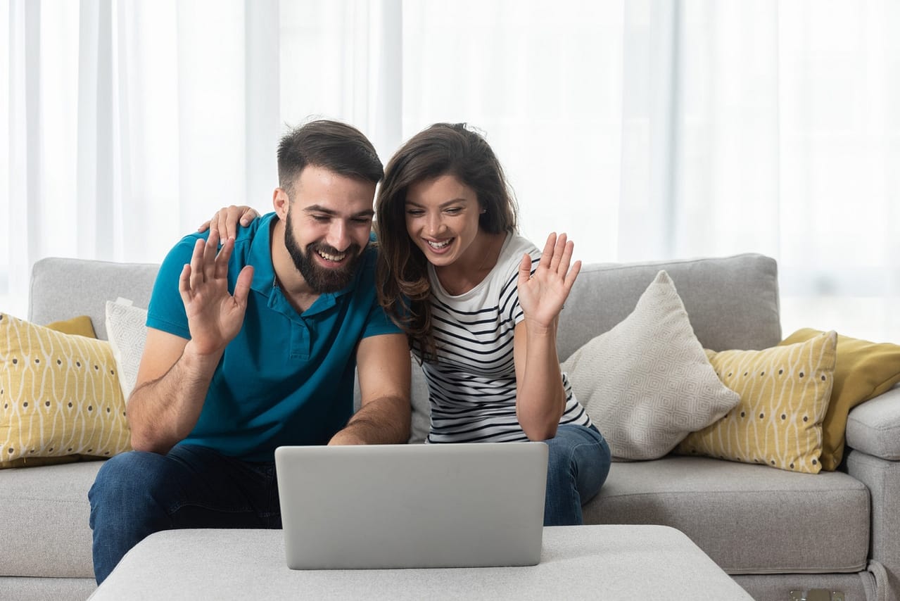 young couple sitting in their living room with laptop waving to
